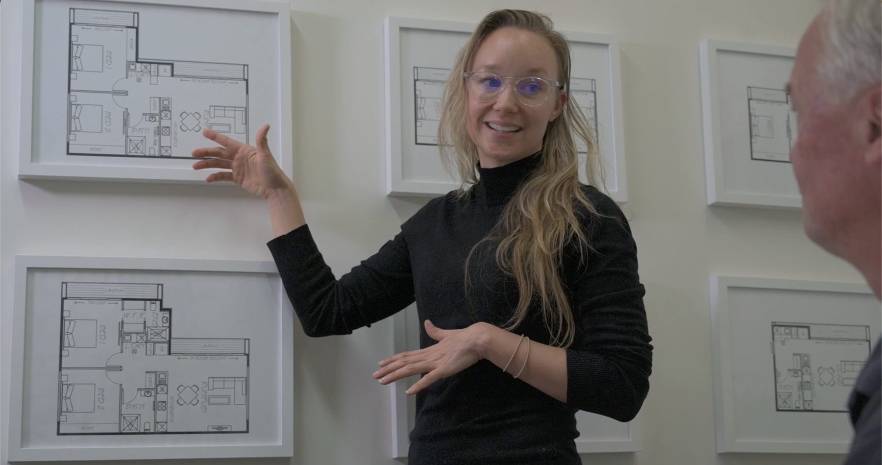 Jacqueline smiling as she walks a client through framed floor plans on the wall