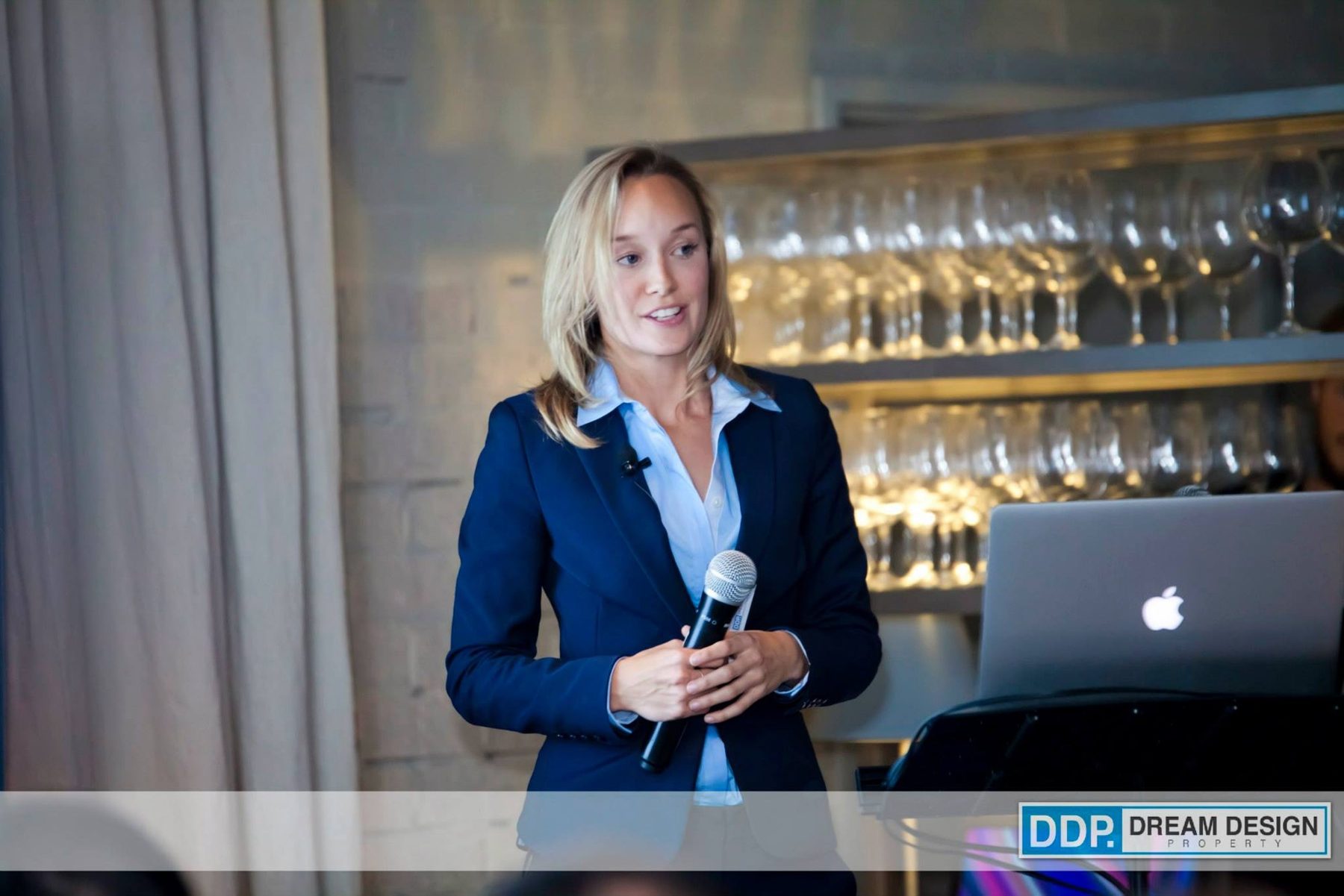 Jacqueline Valk in a navy blazer speaking with a microphone at a corporate event